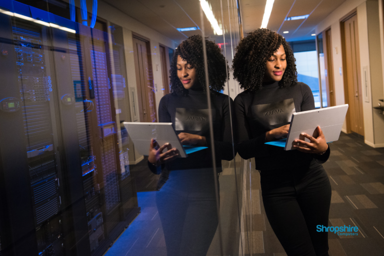 A lady leans against a glass wall, looking at her laptop considering Cyber Essentials.