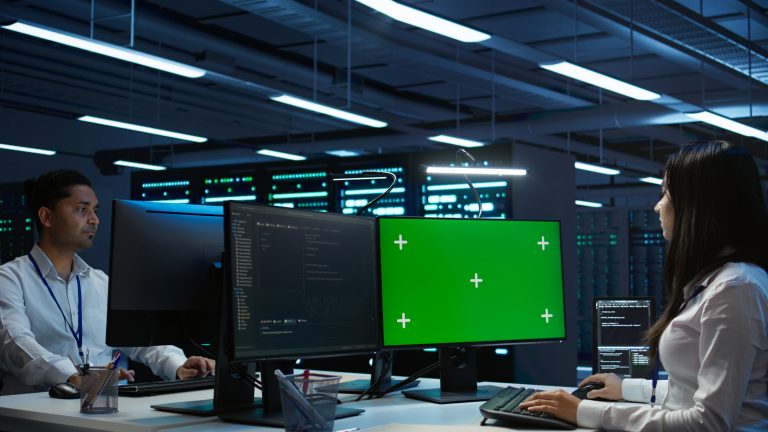 Two IT professionals working at a desk in a dark, high-tech data center with server racks in the background. One monitor shows a green screen mockup with tracking markers.