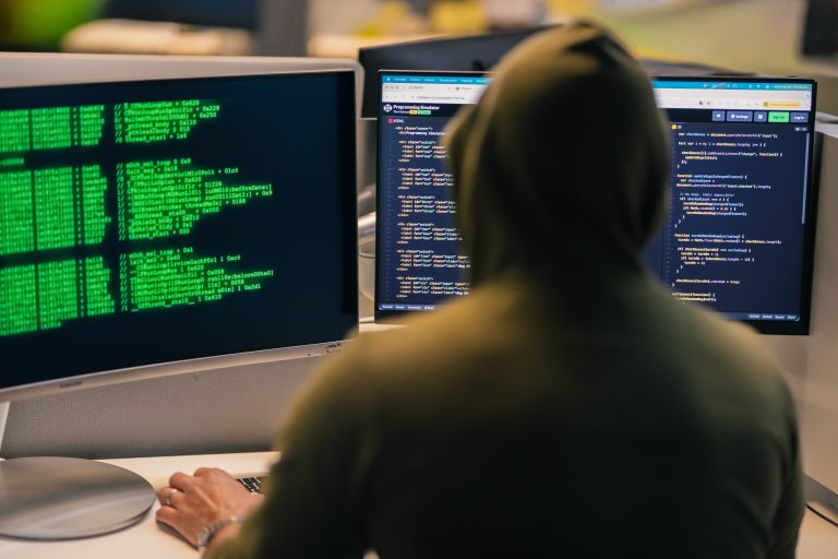 A hooded developer sitting in front of multiple computer monitors displaying green terminal code, HTML, and JavaScript
